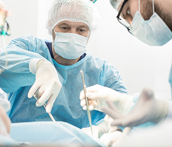 Two surgeons in blue scrubs, masks, and hairnets performing a procedure in a bright operating room