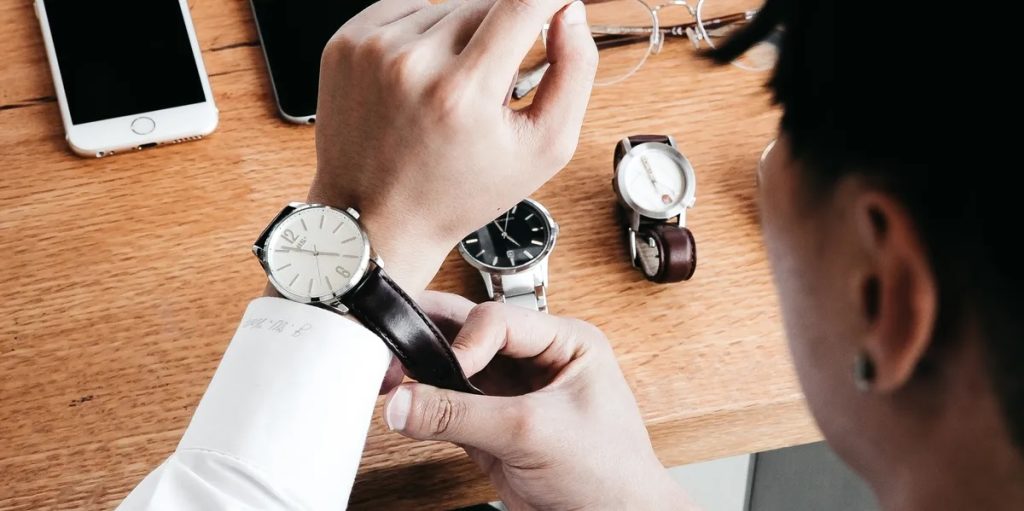 Man fastening a watch on his wrist with phones, watches, and glasses on a desk behind him.